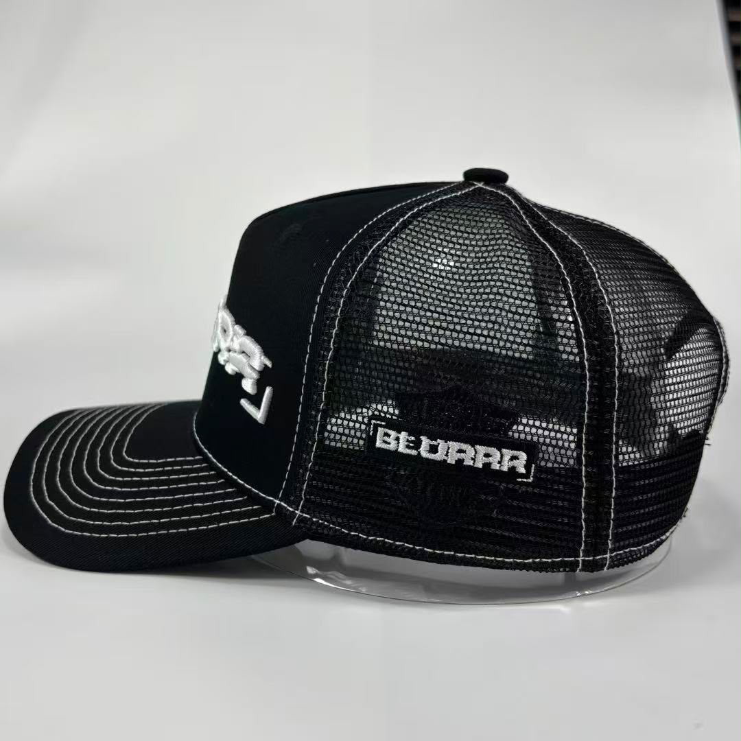 A black trucker-style cap with white contrast stitching and white embroidered logos, displayed on a clear stand against a light gray background.