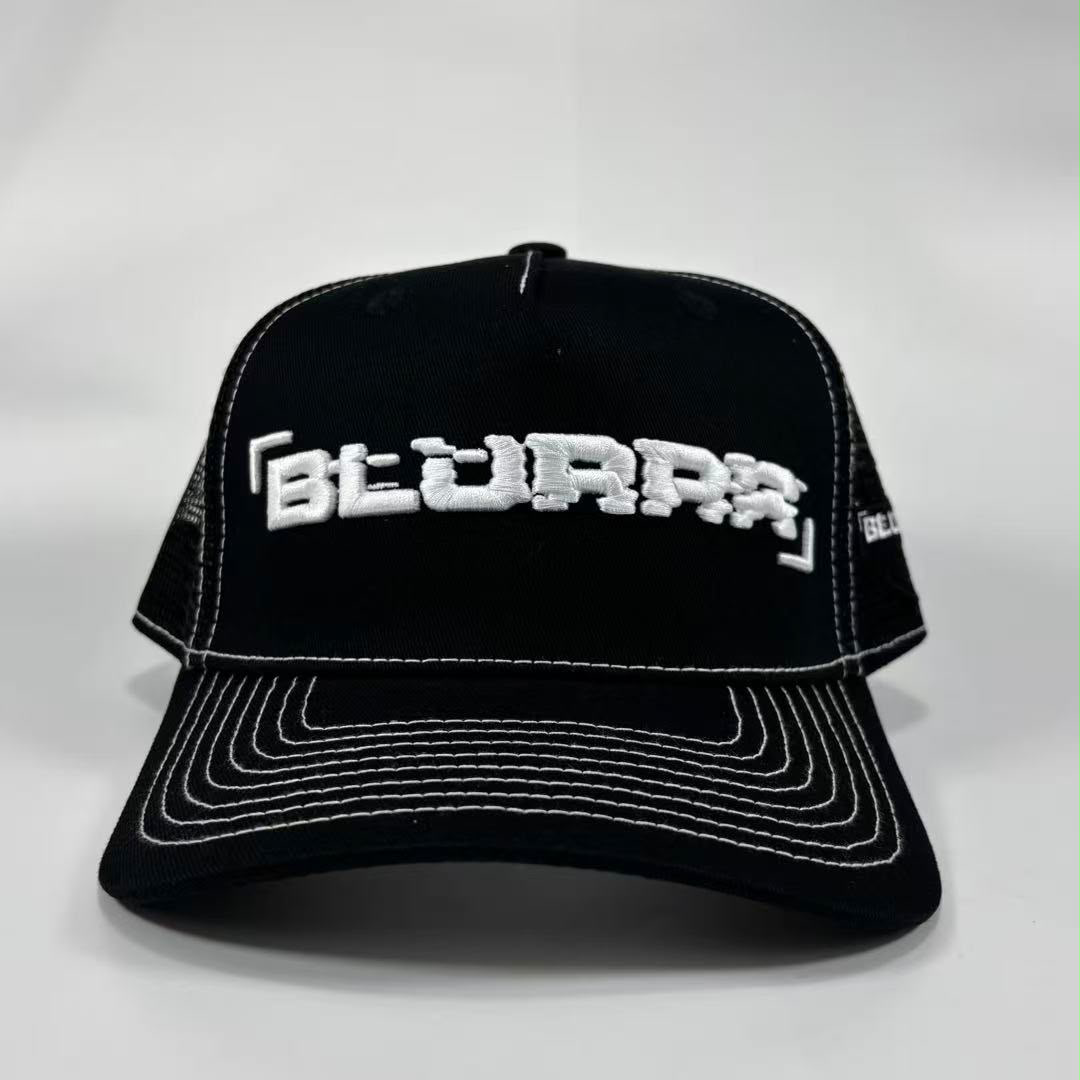 Black trucker-style baseball cap with white contrast stitching and a white embroidered stylised text logo on the front, photographed on a light grey studio background.