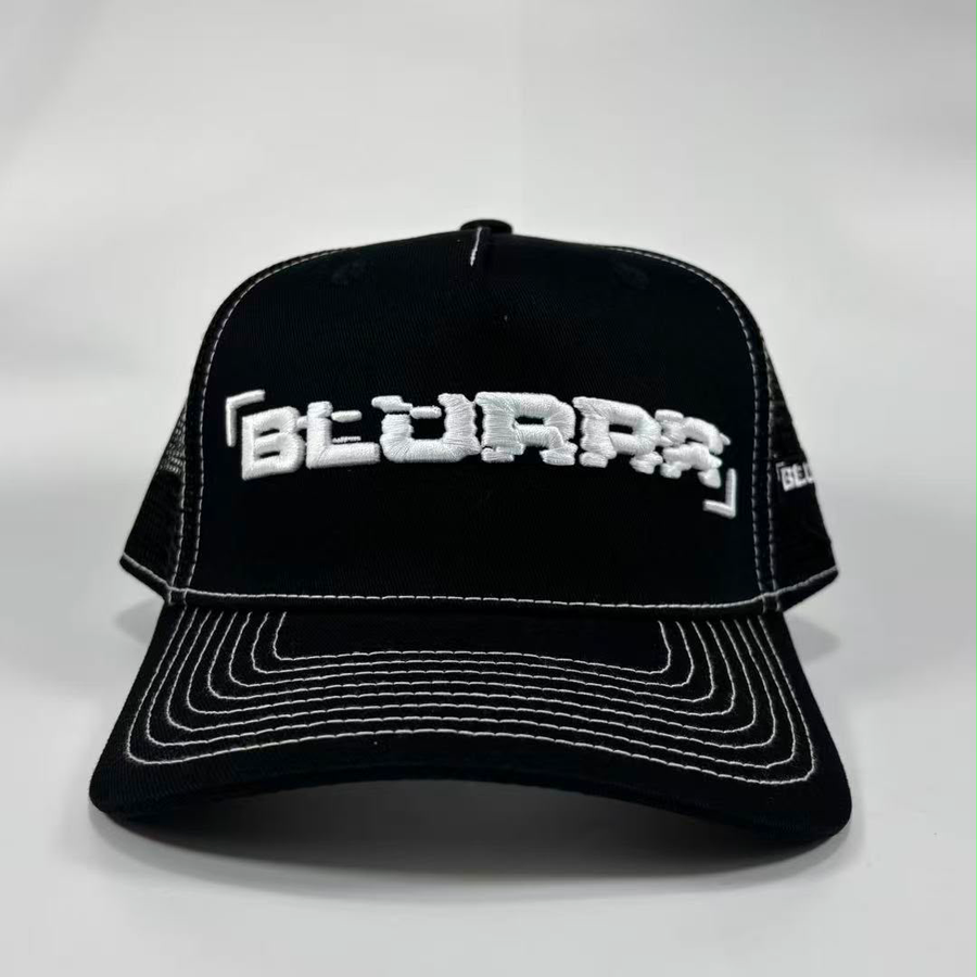 Black trucker-style baseball cap with white contrast stitching and a white embroidered stylised text logo on the front, photographed on a light grey studio background.