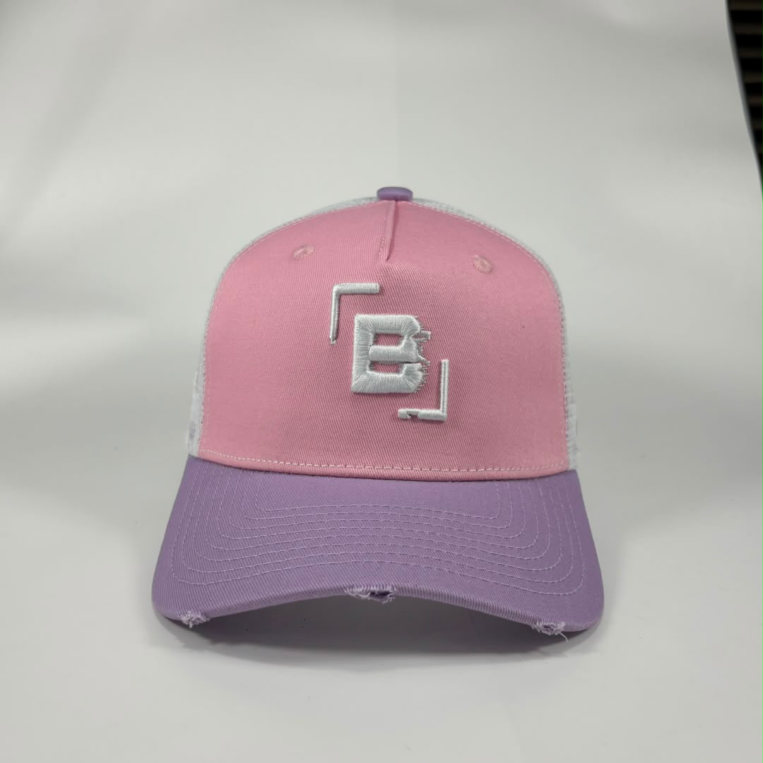 A two-tone trucker-style baseball cap with a pink front panel, lavender-purple brim, and white mesh back. The front panel features a white embroidered geometric 'B' logo.