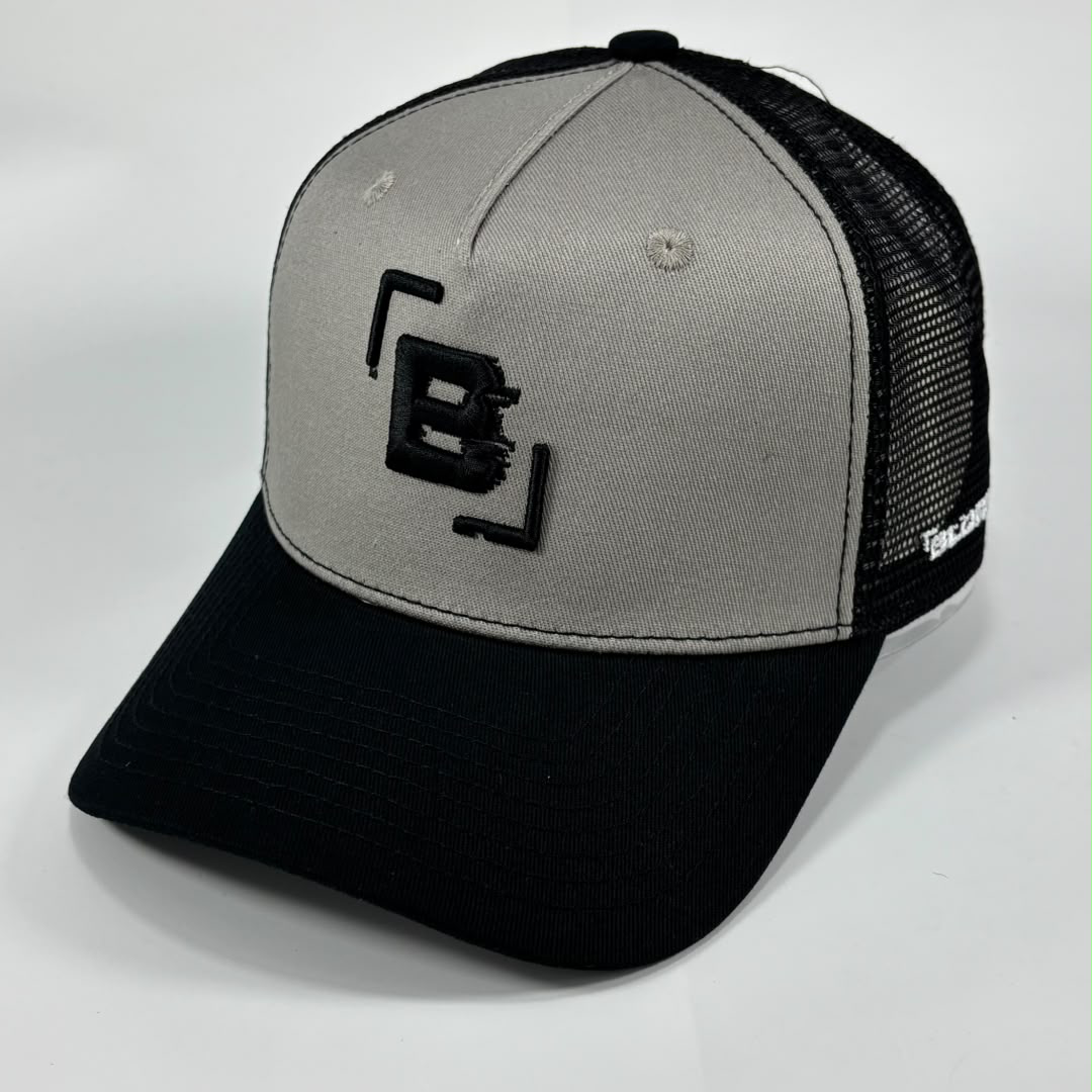 A two-tone trucker hat with a light gray front panel, black mesh back, and black curved brim. The front panel features a black embroidered stylized 'B' logo inside bracket-like borders, and the side of the cap shows the white text 'BLURRR'.