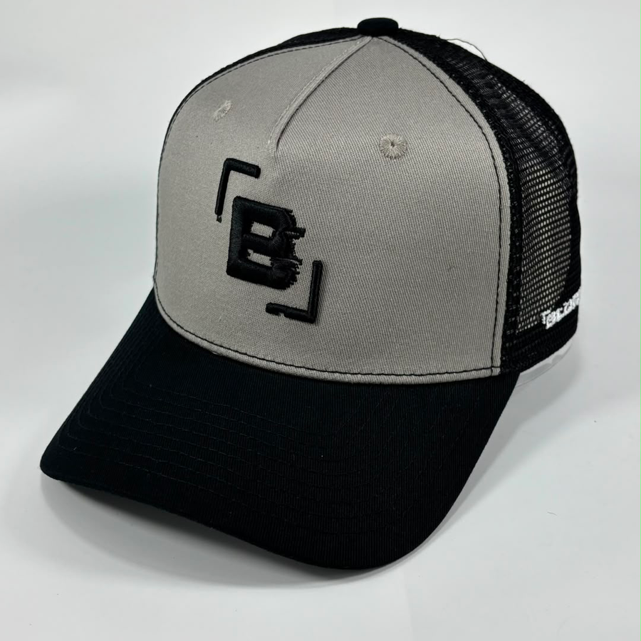 A two-tone trucker hat with a light gray front panel, black mesh back, and black curved brim. The front panel features a black embroidered stylized 'B' logo inside bracket-like borders, and the side of the cap shows the white text 'BLURRR'.