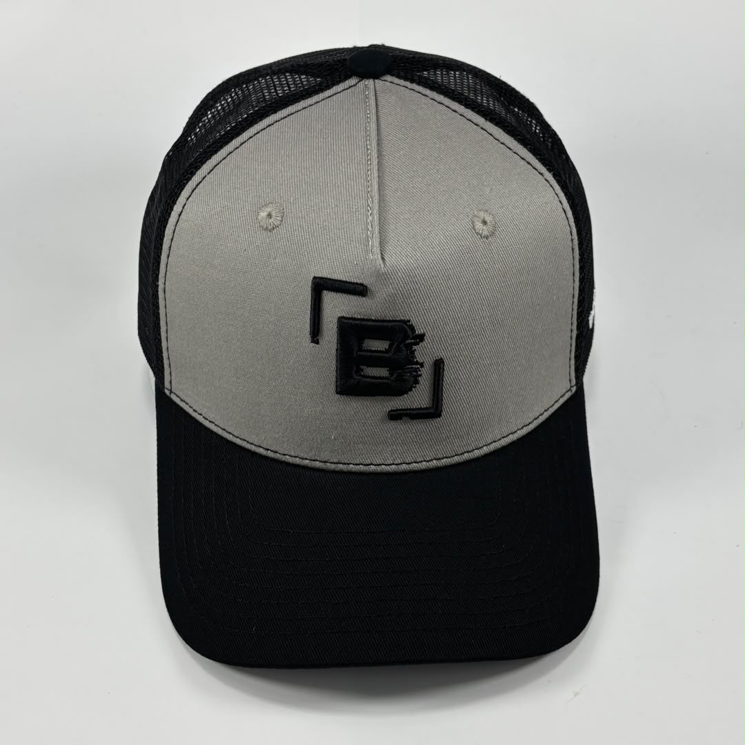A two-tone trucker cap with a light gray front panel and a black brim, mesh back, and black embroidered logo on the front.