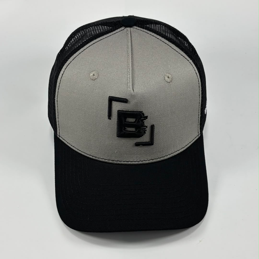 A two-tone trucker cap with a light gray front panel and a black brim, mesh back, and black embroidered logo on the front.