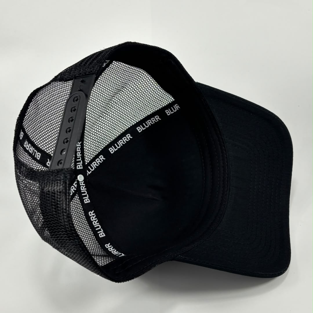 A black trucker-style cap with a black mesh back and a black curved brim, photographed on a light gray background. The mesh band around the back is printed with the brand name 'BLURRR' in white.