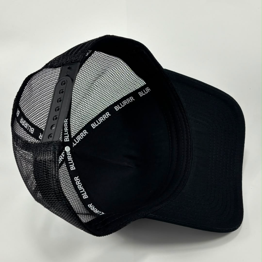 A black trucker-style cap with a black mesh back and a black curved brim, photographed on a light gray background. The mesh band around the back is printed with the brand name 'BLURRR' in white.
