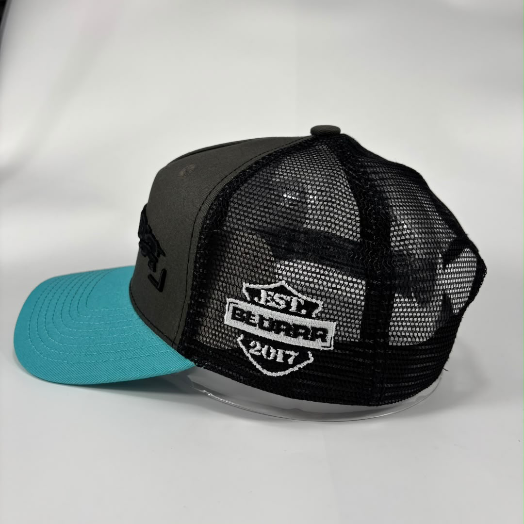 A gray trucker-style baseball cap with a bright turquoise blue brim. The back of the cap features a black mesh back and a white embroidered patch that reads 'EST BLURRR 2017.