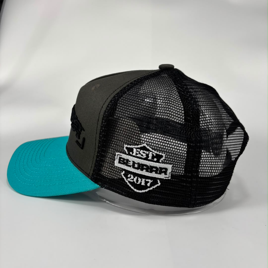 A gray trucker-style baseball cap with a bright turquoise blue brim. The back of the cap features a black mesh back and a white embroidered patch that reads 'EST BLURRR 2017.